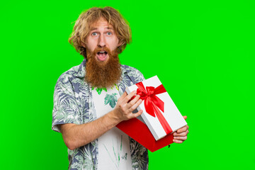 Happy caucasian man opening birthday gift box with red ribbon. Holidays surprise concept. Smiling redhead bearded guy receive great wrapped present celebrating isolated on green chroma key background