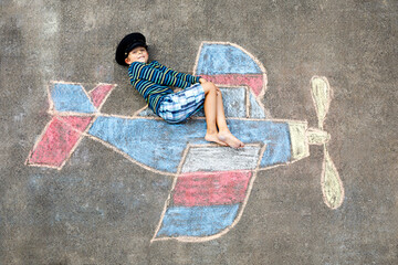 Little kid boy having fun with with airplane picture drawing with colorful chalks on asphalt. Child painting with chalk and crayon and going on vacations or dreaming of pilot profession.