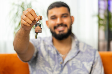 Happy Indian man real estate agent lifting hand showing the keys of new home house apartment, buying or renting property, mortgage loan, investment at modern home room apartment. Guy sits on couch