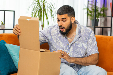 Happy Indian young man unpacking delivery parcel sits at home. Smiling satisfied bearded Arabian guy shopper online shop customer opening cardboard box receive purchase gift by fast postal shipping