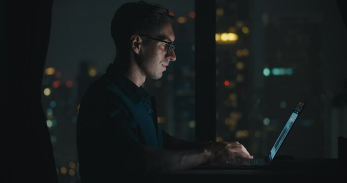 Late night millennial male glasses sits table apartment office works computer apartment. Caucasian mid adult man eyeglasses sitting desk working laptop night time computer screen night city.