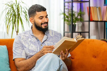 Indian man reading interesting book, turning pages smiling enjoying literature, taking a rest on comfortable couch. Portrait of peaceful cheerful guy relaxing at home apartment living room on sofa