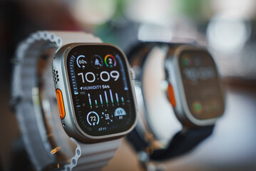 Close up view of two Apple Watches, one with a detailed display showing 10 09 time, humidity, temperature, and activity rings. Silver and orange accents in focus at a retail setting.