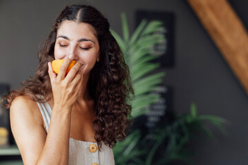 A young attractive woman closes her eyes and sniffs half an orange. A curly-haired smiling girl is...