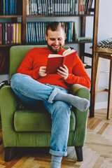 Cheerful caucasian young man laughing on funny story enjoying free time at cozy home interior near bookcase in living room,positive hipster guy concentrated on reading interesting book in armchair