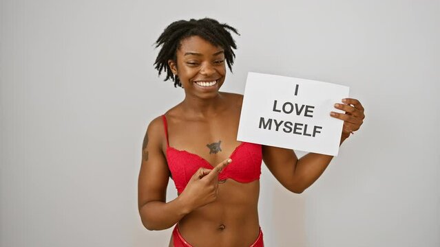 Cheerful young black woman with dreadlocks confidently pointing and smiling at 'i love my body' text she's presenting, optimistically marking an idea of self-love on isolated white background.