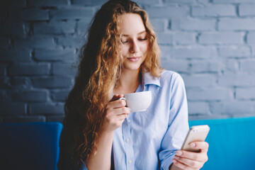 Charming female blogger enjoying tea time in cafeteria and sending text messages to followers via...
