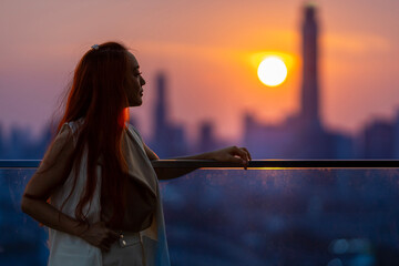 Woman looking and enjoying the sunset view from balcony with the sun setting behind skyscraper in busy urban downtown with loneliness for solitude, loneliness and dreaming of freedom lifestyle