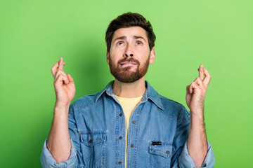 Photo of unsure handsome guy wear denim jacket fingers crossed looking up empty space isolated...