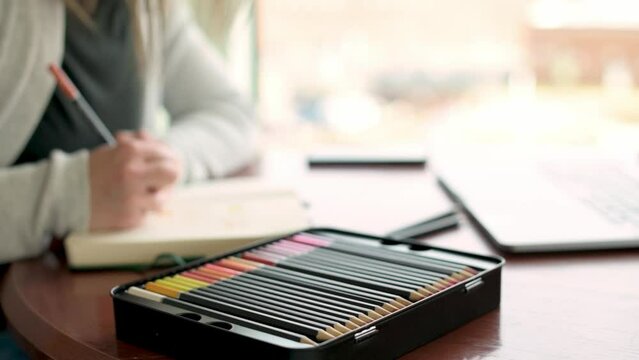 artist drawing in a notebook at a well-lit desk, with a sharp focus on a tin box of colored pencils in the foreground. The setting promotes a creative and peaceful atmosphere ideal for art and design.