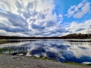 Spring's Canvas: A tranquil lake mirrors the drama of a spring sky, with clouds painted in soft grays and whites above a quiet, awakening forest lining the shore.