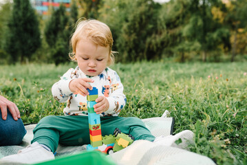 Little cute toddler boy playing small car toys in garden. Child 2 year old plays constructor game sitting in green grass on blanket, walking in park sunny summer day. Outdoors creative activities.