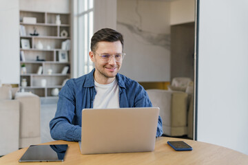 A content young professional engaging with his laptop at a wooden table, in a well-lit, elegant living space, conveys focus and satisfaction.