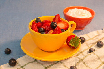 A yellow cup is filled with strawberries and blueberries, with a cup of whipped cream in the background