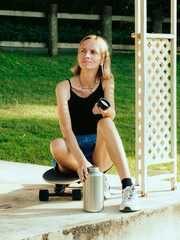 Woman feels thirsty, sitting with skateboard and water bottle at hot summer day.