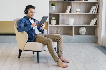 A young professional comfortably sits barefoot in an armchair, deeply engaged with a tablet and listening to music through black headphones, surrounded by a stylishly minimalistic decor.