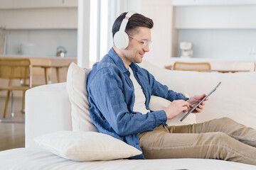 A relaxed young professional enjoys music through white headphones while using a tablet on a comfortable sofa, in a well-lit, modern living space.