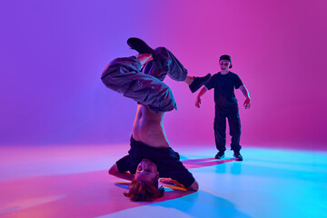 Two young boys, dancers participate in street dance battle in mixed neon light against vibrant gradient background. Concept of sport and hobby, music, fashion and art, movement. Ad