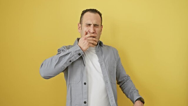 Attractive young hispanic man in shirt, yawning from boredom and sleepiness, hand over mouth. tired, standing isolated on yellow background, lazing around in morning's yawning fatigue