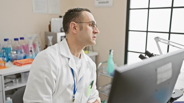 Focused hispanic man in lab coat analyzing data on a computer in a laboratory setting.