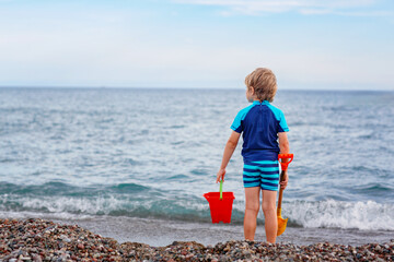 Adorable little blond kid boy standing on lonely ocean beach. Child playing with bucket and shovel...