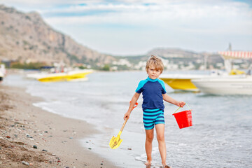 Adorable little blond kid boy walking on ocean beach. Child playing with bucket and shovel and...