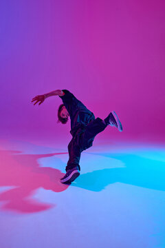 Junior street dancer in dynamic pose dressed in all black in mixed neon light against vibrant gradient background. Concept of sport and hobby, music, fashion and art, movement. Ad