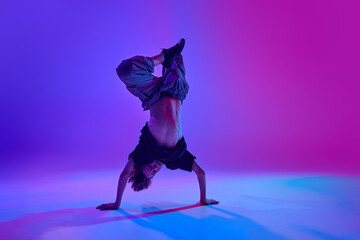 Stylish dressed young dancer, guy performing headstand in motion in mixed neon light against...
