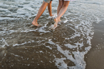 Down view of bare feet on sand beach. Couple in love walking barefoot on water with big waves on seashore. Man and woman walk on sea. Spending time together. Bottom view on legs. Back view.