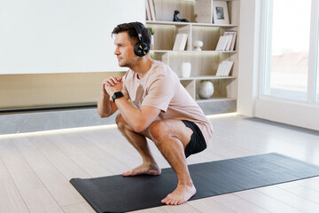 A focused man wearing headphones performs a squat on a yoga mat in a bright, modern room, enhancing his fitness routine.
