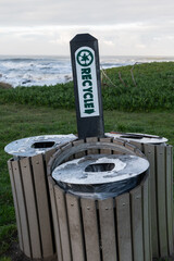 Multiple recycling bins in a grassy field on the shore of the Pacific Ocean.