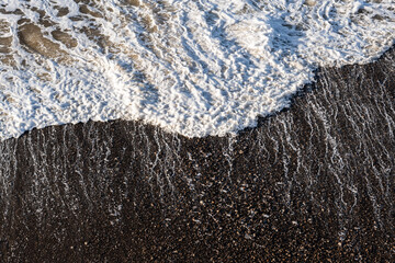 Detail of a white, foamy wave arriving on the shore of a black, rocky beach.
