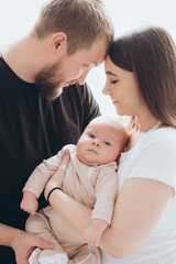 mom and dad with a baby in their arms on a white isolated window background, parents hugging and kissing a newborn baby at home by the window, a happy family