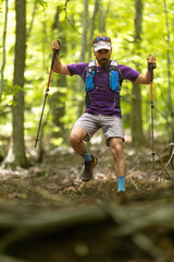 A man wearing a purple shirt and gray shorts is holding two ski poles and jumping over a log