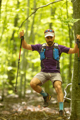 A man wearing a purple shirt and gray shorts is jumping over a tree. He is holding two poles in his...