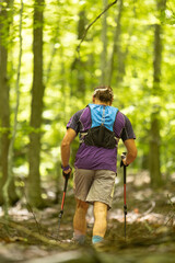A man is walking through a forest with a backpack on. He is wearing a purple shirt and shorts