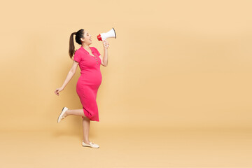 Portrait of Happy Asian pregnant woman standing and holding megaphone isolated on brown background,...