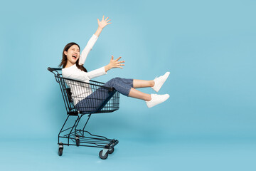 Happy young Asian woman sitting inside of shopping trolley isolated on blue sky background, Wow and...