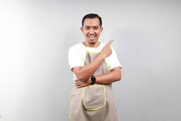 Happy asian barista pointing finger left and smiling, wearing brown apron uniform, standing against white background. Copy space
