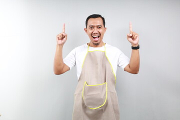 Surprised asian male waiter in brown apron showing logo or advertisement, pointing fingers up at...