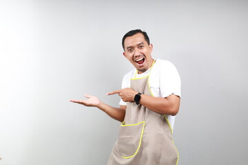 Excited asian man with brown apron showing something with his hand isolated on gray background