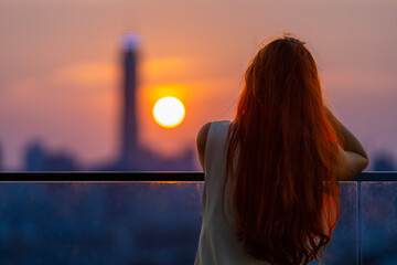 Woman looking and enjoying the sunset view from balcony with the sun setting behind skyscraper in...