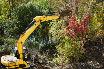 Excavator bucket removes green vegetation, clearing the construction site from green vegetation,...