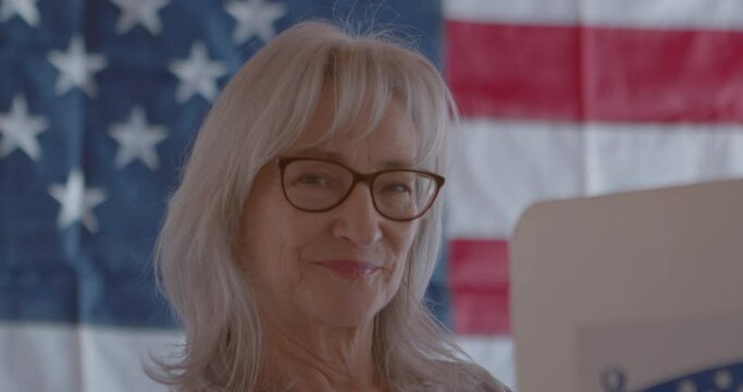 Close-up of a mature woman voter smiling  and voting behind a polling booth with an American flag in the background