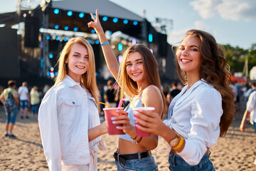 Group of happy females cheers with cocktails at a lively beach music festival. Smiling friends enjoying party, summer vibe, coastline backdrop. Girls toast drinks, celebrate, dance to live band.