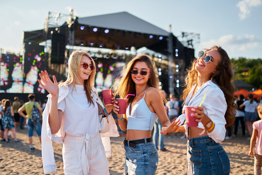 Girls enjoying summer music festival on beach, holding drinks, dancing in sunshine. Group of young women celebrate, having fun at outdoor concert, vibrant fest scene with crowd, stage in background.