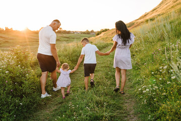 Dad and mom holding hands baby daughter, son walk in mountains at sunset. Family spending time together on a summer day. Children with parents happiness walking in green grass in field. Back view