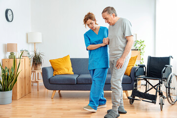 Young caregiver assisting senior man in wheelchair walking indoors - Home health care service and...
