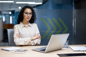 A professional Hispanic businesswoman poses confidently at her desk in a contemporary office...