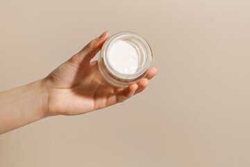 A female hand holds a glass jar with white face cream on a beige background. The concept of beauty, skin care, cosmetology.